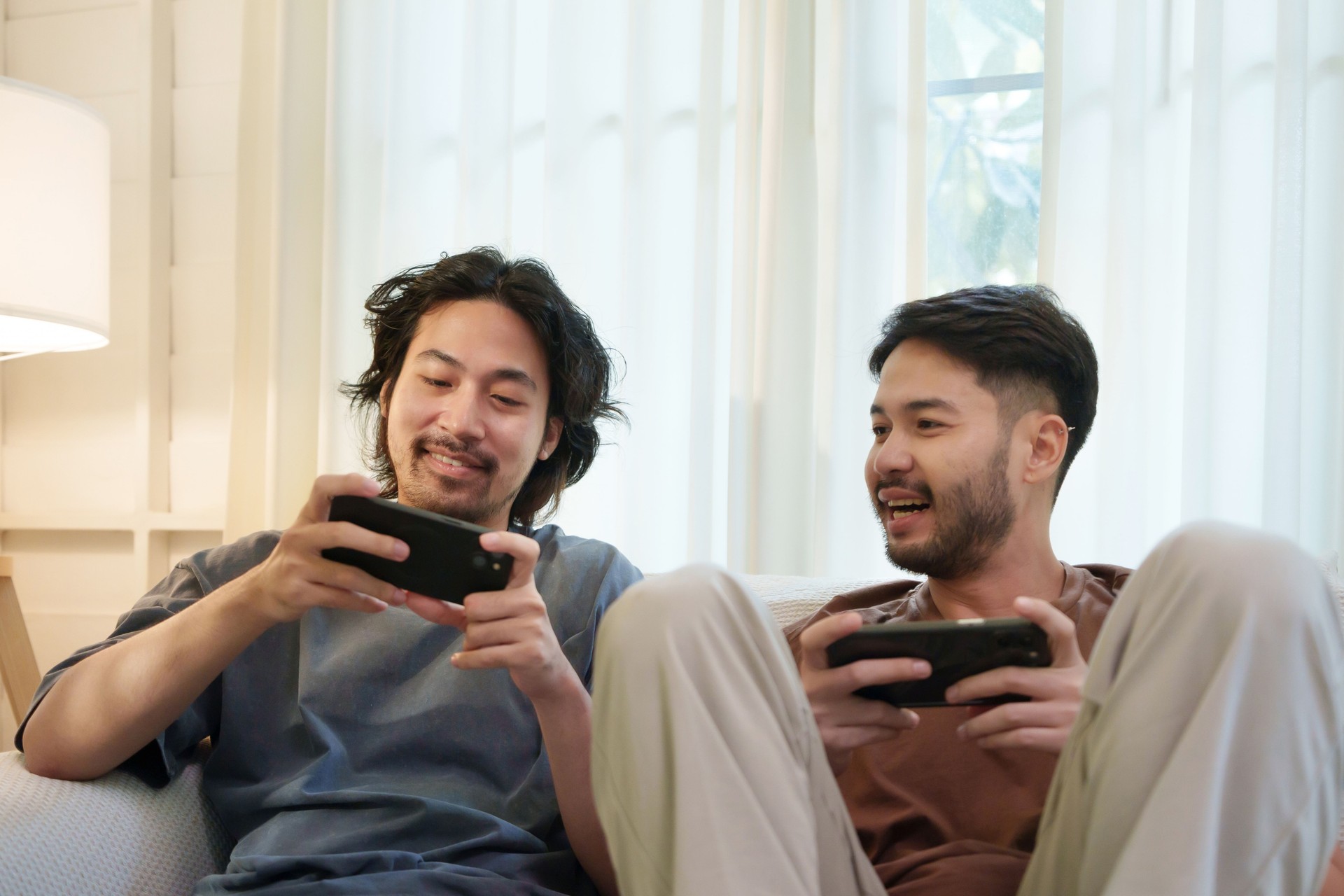 Relaxed Two Asian men playing mobile games together on a sofa. Two friends smiling and enjoying a friendly competition on smartphone.
